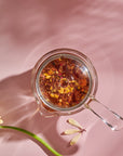 A glass Tea in Hand Teapot from The Best of Magic Hour Sampler Set filled with herbal tea, viewed from above next to a yellow flower on a pink surface.