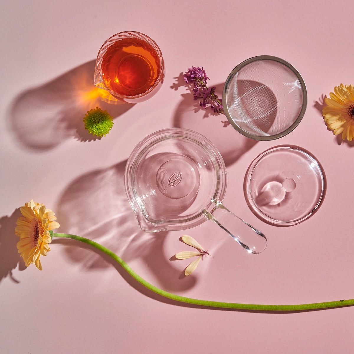 Aerial view of various borosilicate glass objects on a pink surface. The setup includes an Espresso Parts Tea-in-Hand: The Perfect Steep Side-pour Ceremonial Teapot, an empty round dish, a glass with orange liquid, and a small clear dish. The arrangement is complemented by loose leaf teas and features a green stem with a yellow flower and small purple flowers.