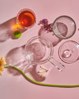 Aerial view of various borosilicate glass objects on a pink surface. The setup includes an Espresso Parts Tea-in-Hand: The Perfect Steep Side-pour Ceremonial Teapot, an empty round dish, a glass with orange liquid, and a small clear dish. The arrangement is complemented by loose leaf teas and features a green stem with a yellow flower and small purple flowers.