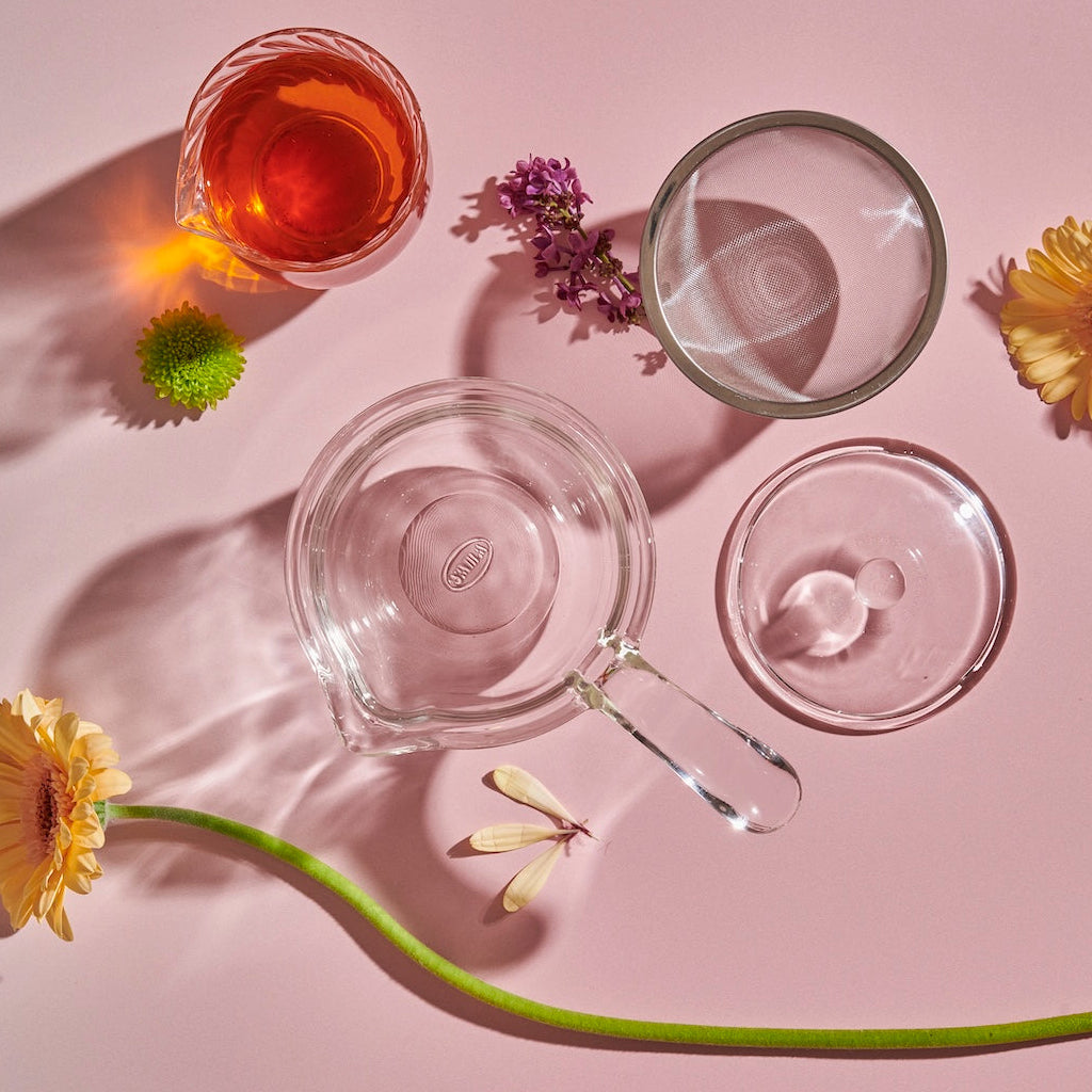 A Tea-in-Hand: The Perfect Steep Side-pour Ceremonial Teapot, two cups (one with a reddish liquid), flowers, and a glass stirrer are arranged artistically on a pink background. The shadows cast by the objects and flowers create a visually appealing composition, perfect for enjoying loose leaf teas.