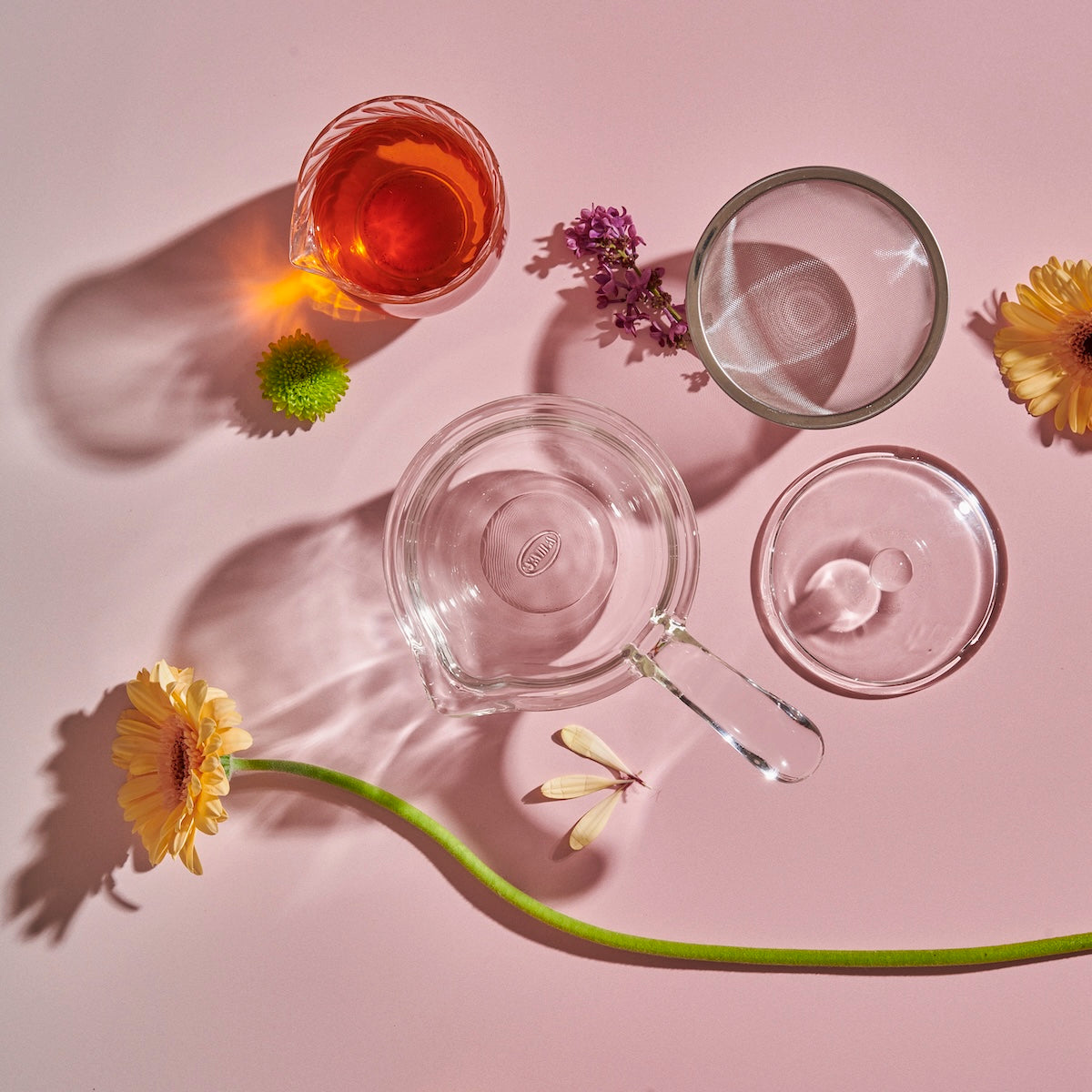 A Tea-in-Hand: The Perfect Steep Side-pour Ceremonial Teapot by Espresso Parts, two cups (one with a reddish liquid), flowers, and a glass stirrer are arranged artistically on a pink background. The shadows cast by the objects and flowers create a visually appealing composition, perfect for enjoying loose leaf teas.