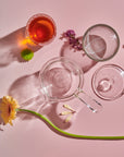 A Tea-in-Hand: The Perfect Steep Side-pour Ceremonial Teapot by Espresso Parts, two cups (one with a reddish liquid), flowers, and a glass stirrer are arranged artistically on a pink background. The shadows cast by the objects and flowers create a visually appealing composition, perfect for enjoying loose leaf teas.