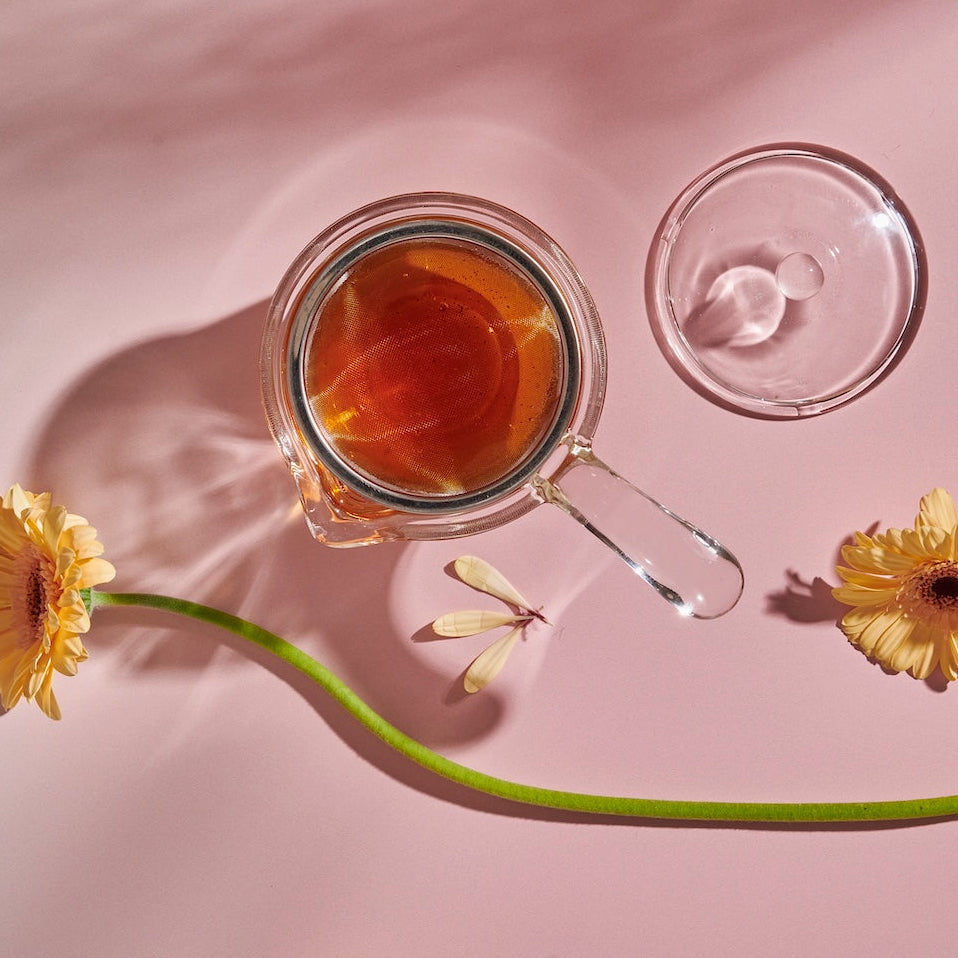A glass cup filled with loose leaf tea sits on a light pink surface next to a Tea-in-Hand: The Perfect Steep Side-pour Ceremonial Teapot made of borosilicate glass by Espresso Parts. Two yellow daisies and their shadows are visible, one lying horizontally across the table and the other at an angle, adding a touch of nature to the setting.