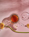 A glass cup filled with loose leaf tea sits on a light pink surface next to a Tea-in-Hand: The Perfect Steep Side-pour Ceremonial Teapot made of borosilicate glass by Espresso Parts. Two yellow daisies and their shadows are visible, one lying horizontally across the table and the other at an angle, adding a touch of nature to the setting.