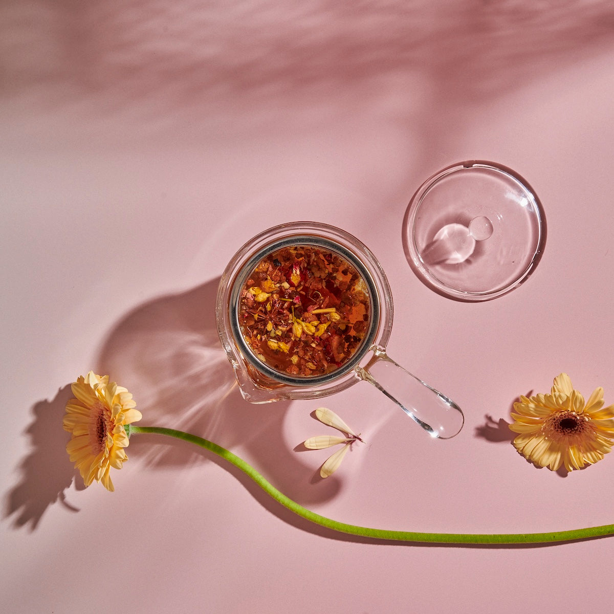A top view of a borosilicate glass Espresso Parts Tea-in-Hand: The Perfect Steep Side-pour Ceremonial Teapot containing an infusion of loose leaf teas, positioned against a pale pink background. The teapot's lid is placed beside it. Two yellow daisies and a delicate dragonfly decoratively accompany the scene.