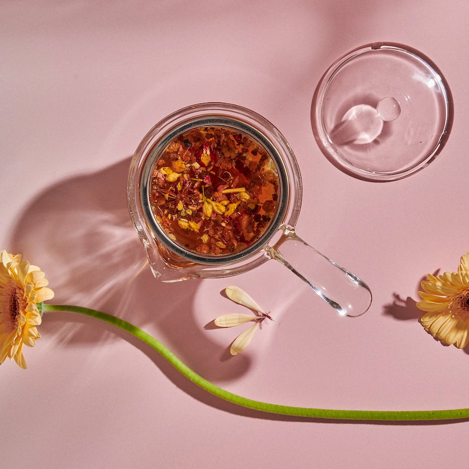 A top view of a borosilicate glass Espresso Parts Tea-in-Hand: The Perfect Steep Side-pour Ceremonial Teapot containing an infusion of loose leaf teas, positioned against a pale pink background. The teapot's lid is placed beside it. Two yellow daisies and a delicate dragonfly decoratively accompany the scene.