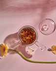 A top view of a borosilicate glass Espresso Parts Tea-in-Hand: The Perfect Steep Side-pour Ceremonial Teapot containing an infusion of loose leaf teas, positioned against a pale pink background. The teapot's lid is placed beside it. Two yellow daisies and a delicate dragonfly decoratively accompany the scene.