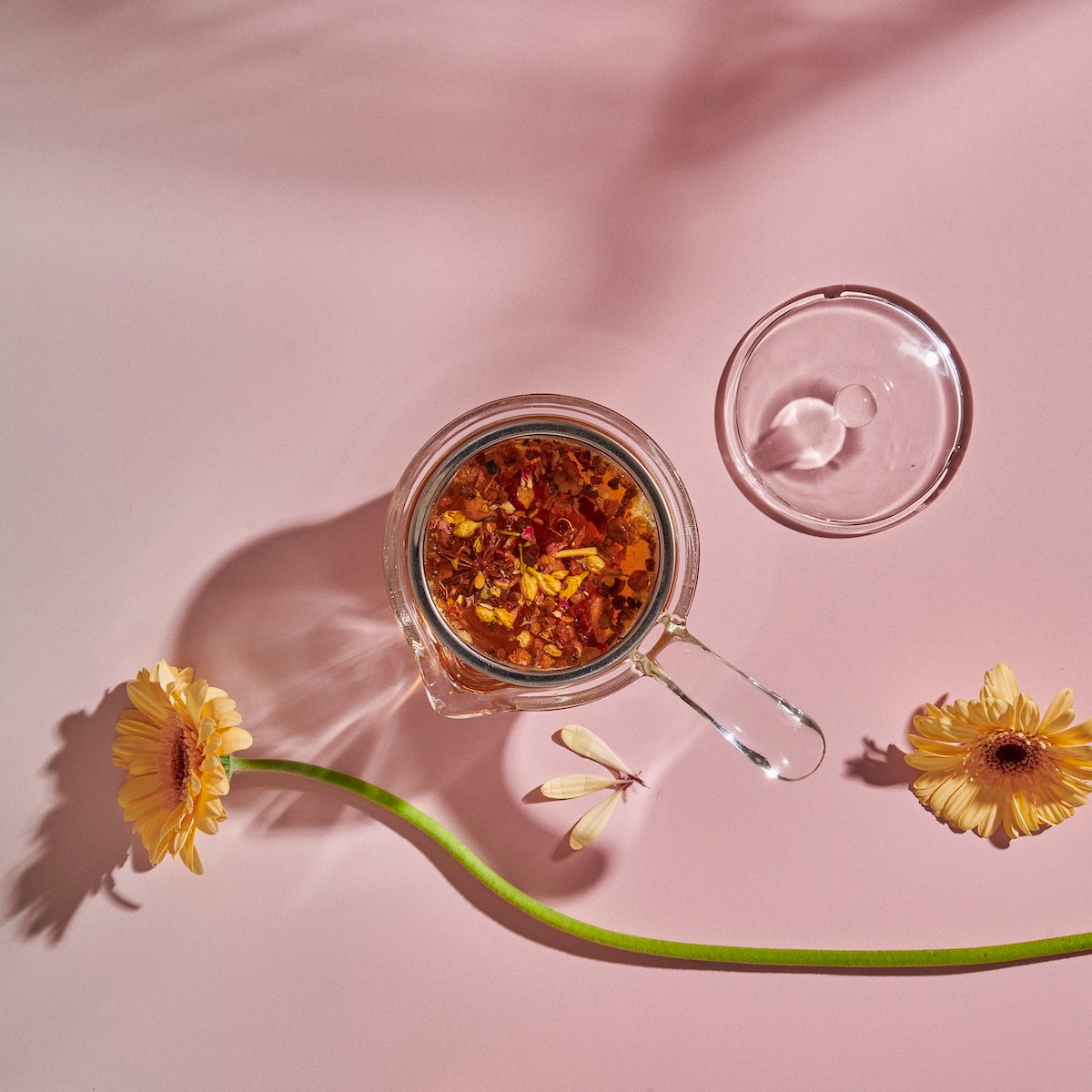 A side-handled Tea-in-Hand: The Perfect Steep Side-pour Ceremonial Teapot by Espresso Parts made of borosilicate glass with an infusion of herbs and flower petals sits on a pink surface. The teapot lid is placed beside it. Two yellow gerbera daisies and a long green stem are arranged around the teapot, adding a touch of natural beauty perfect for loose leaf teas.