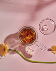 A side-handled Tea-in-Hand: The Perfect Steep Side-pour Ceremonial Teapot by Espresso Parts made of borosilicate glass with an infusion of herbs and flower petals sits on a pink surface. The teapot lid is placed beside it. Two yellow gerbera daisies and a long green stem are arranged around the teapot, adding a touch of natural beauty perfect for loose leaf teas.