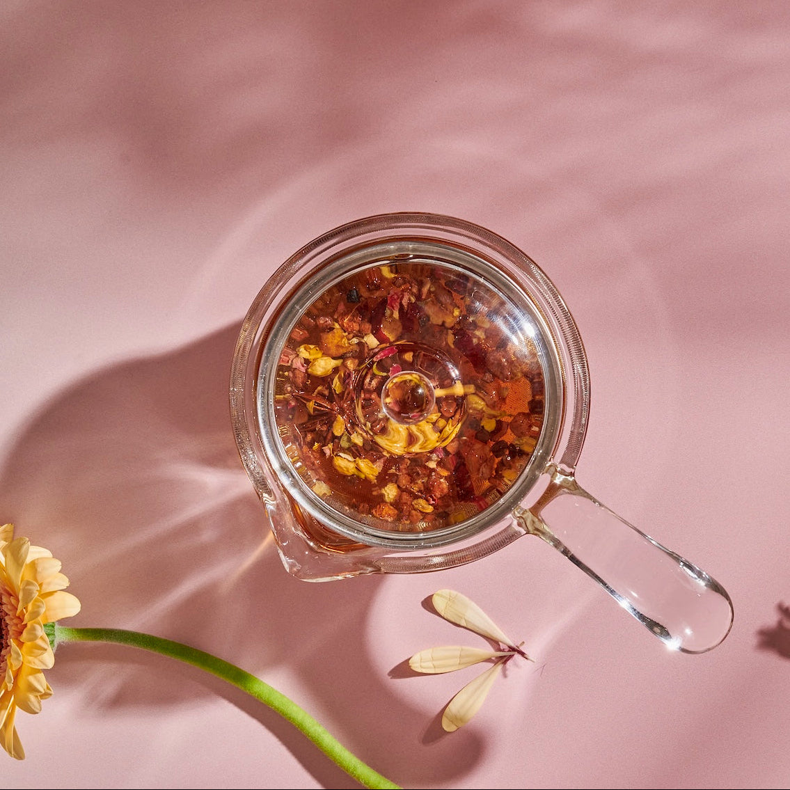A **Tea-in-Hand: The Perfect Steep Side-pour Ceremonial Teapot** by **Espresso Parts** made of borosilicate glass filled with floral loose leaf teas is placed on a pink surface with a daisy and a decorative model of a dragonfly nearby. The shadows of the daisy and dragonfly are visible, adding a whimsical feel to the scene.