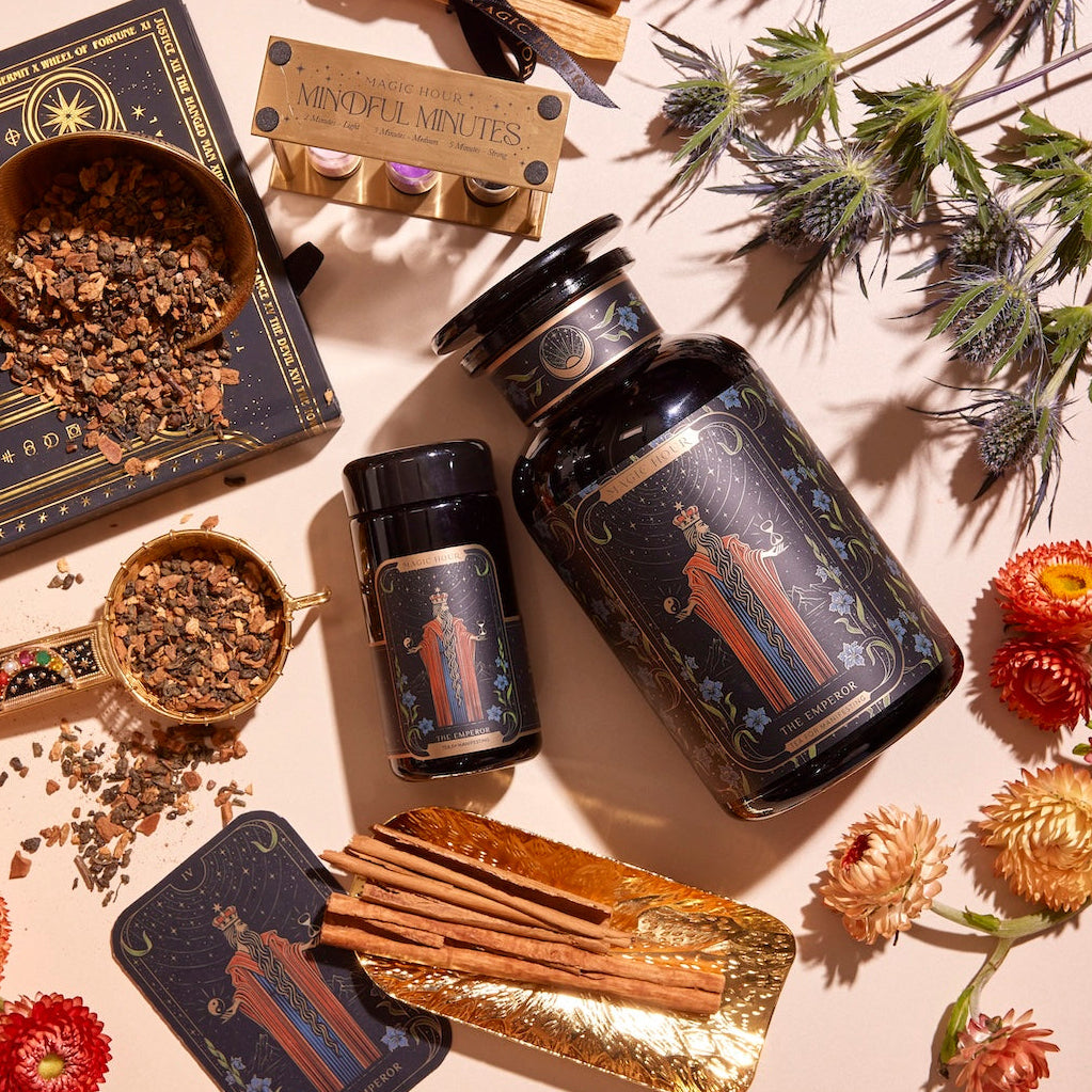 A flat lay of wellness products including jars of herbs, The Emperor: Green Chaga Chai with Lion's Mane and Reishi for immune support, a tin containing incense sticks, cinnamon sticks, dried flowers, and a box of mindful minutes cards. The items are beautifully arranged on a white surface with decorative gold and black paper underneath some pieces.