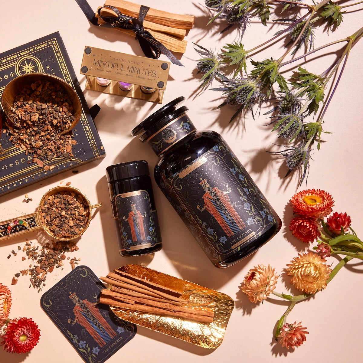 A flat lay of wellness products including jars of herbs, The Emperor: Green Chaga Chai with Lion's Mane and Reishi for immune support, a tin containing incense sticks, cinnamon sticks, dried flowers, and a box of mindful minutes cards. The items are beautifully arranged on a white surface with decorative gold and black paper underneath some pieces.