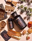 A flat lay of wellness products including jars of herbs, The Emperor: Green Chaga Chai with Lion's Mane and Reishi for immune support, a tin containing incense sticks, cinnamon sticks, dried flowers, and a box of mindful minutes cards. The items are beautifully arranged on a white surface with decorative gold and black paper underneath some pieces.