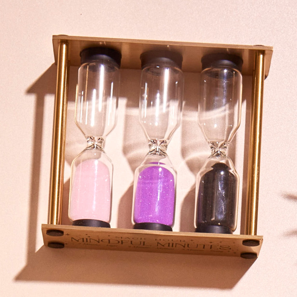 The "Mindful Minutes" hourglass set features three sand timers filled with pink, purple, and black sand, all mounted on a wooden frame, designed to help you time activities thoughtfully.