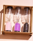 The "Mindful Minutes" hourglass set features three sand timers filled with pink, purple, and black sand, all mounted on a wooden frame, designed to help you time activities thoughtfully.