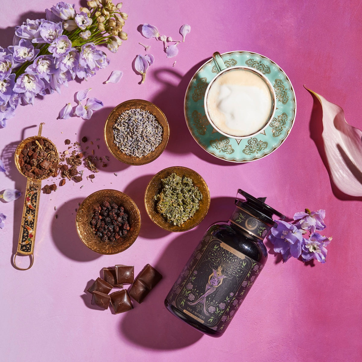 A flat lay of The Empress: Lavender Currant Shatavari Cocoa Tea of Nurturing Creativity by Magic Hour in an ornate cup and saucer, surrounded by dried flowers, organic herbs, chocolate pieces, and a decorative jar on a pink and purple gradient background. A sprig of fresh purple flowers and a white calla lily are also in the scene.