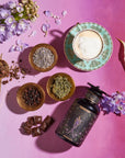 A flat lay of The Empress: Lavender Currant Shatavari Cocoa Tea of Nurturing Creativity by Magic Hour in an ornate cup and saucer, surrounded by dried flowers, organic herbs, chocolate pieces, and a decorative jar on a pink and purple gradient background. A sprig of fresh purple flowers and a white calla lily are also in the scene.