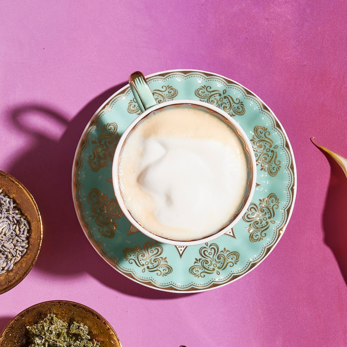 A cup of cappuccino with frothy milk on top is placed on a decorative turquoise saucer with intricate patterns. The saucer rests on a pink surface. Surrounding the cup are small bowls containing organic herbs, creating an inviting atmosphere for a Magic Hour: The Empress: Lavender Currant Shatavari Cocoa Tea of Nurturing Creativity experience.