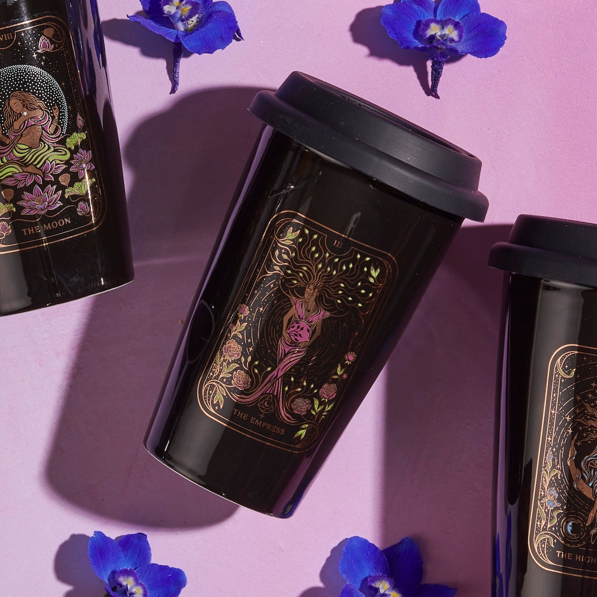 Three black travel mugs featuring tarot card designs—&quot;The Moon,&quot; &quot;The Empress,&quot; and &quot;The High Priestess&quot;—are arranged on a pink surface. Surrounded by scattered purple flowers, these mugs set the perfect scene for enjoying your The Queen of Cups - Tarot To Go! or Magic Hour in style.
