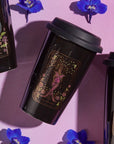 Three black travel mugs featuring tarot card designs—"The Moon," "The Empress," and "The High Priestess"—are arranged on a pink surface. Surrounded by scattered purple flowers, these mugs set the perfect scene for enjoying your The Queen of Cups - Tarot To Go! or Magic Hour in style.