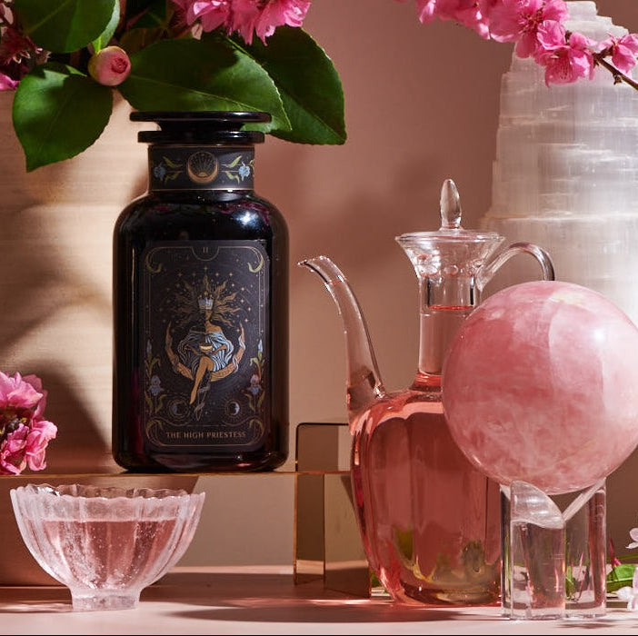 A decorative arrangement featuring a dark glass bottle with ornate design, a clear glass jug of Magic Hour's The High Priestess: Wisdom Tea for Powerful Dreams & Illuminated Insights, a pink crystal sphere on a stand, a white crystal tower, a black crystal obelisk, and a bouquet of pink flowers in a vase, all on a pink surface.