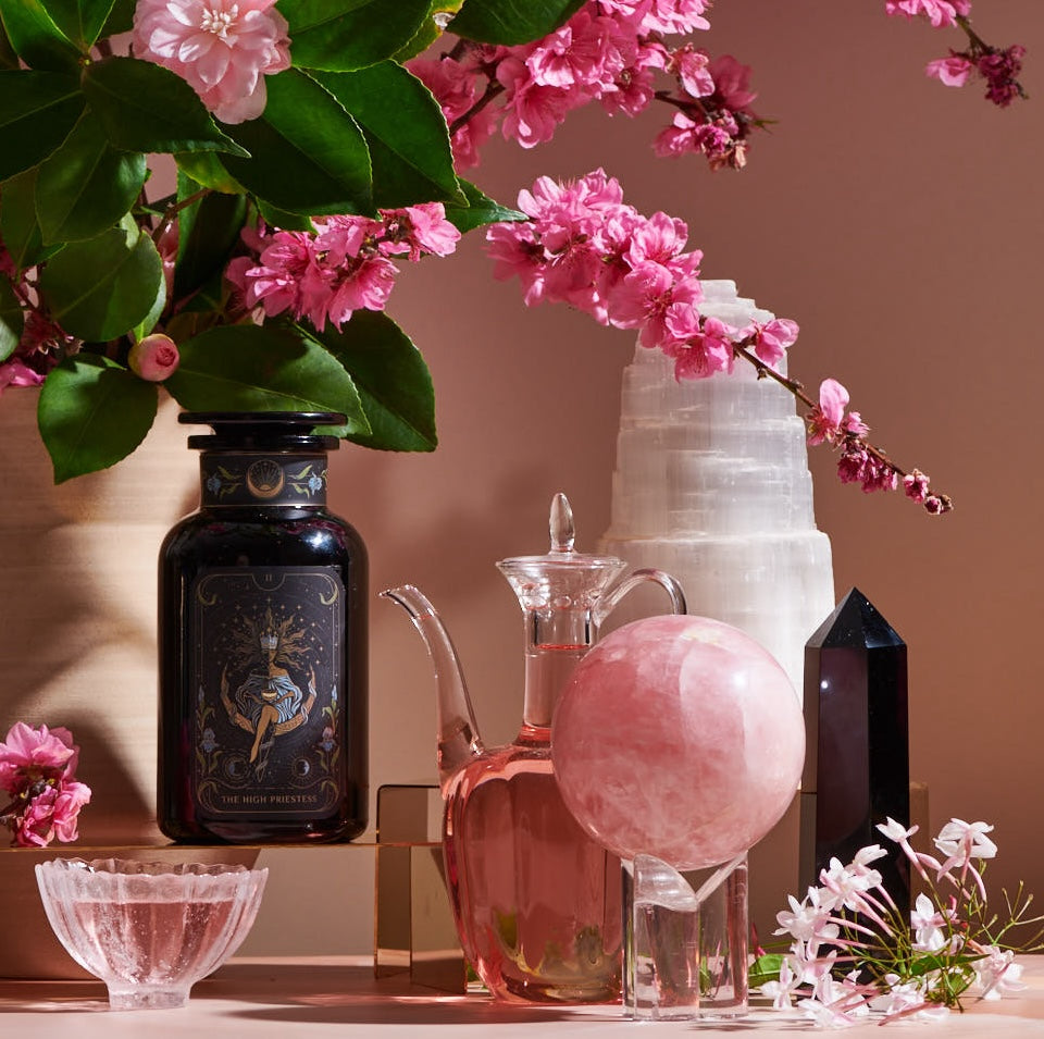 A decorative arrangement featuring a dark glass bottle with ornate design, a clear glass jug of Magic Hour&#39;s The High Priestess: Wisdom Tea for Powerful Dreams &amp; Illuminated Insights, a pink crystal sphere on a stand, a white crystal tower, a black crystal obelisk, and a bouquet of pink flowers in a vase, all on a pink surface.