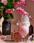 A decorative arrangement featuring a dark glass bottle with ornate design, a clear glass jug of Magic Hour's The High Priestess: Wisdom Tea for Powerful Dreams & Illuminated Insights, a pink crystal sphere on a stand, a white crystal tower, a black crystal obelisk, and a bouquet of pink flowers in a vase, all on a pink surface.