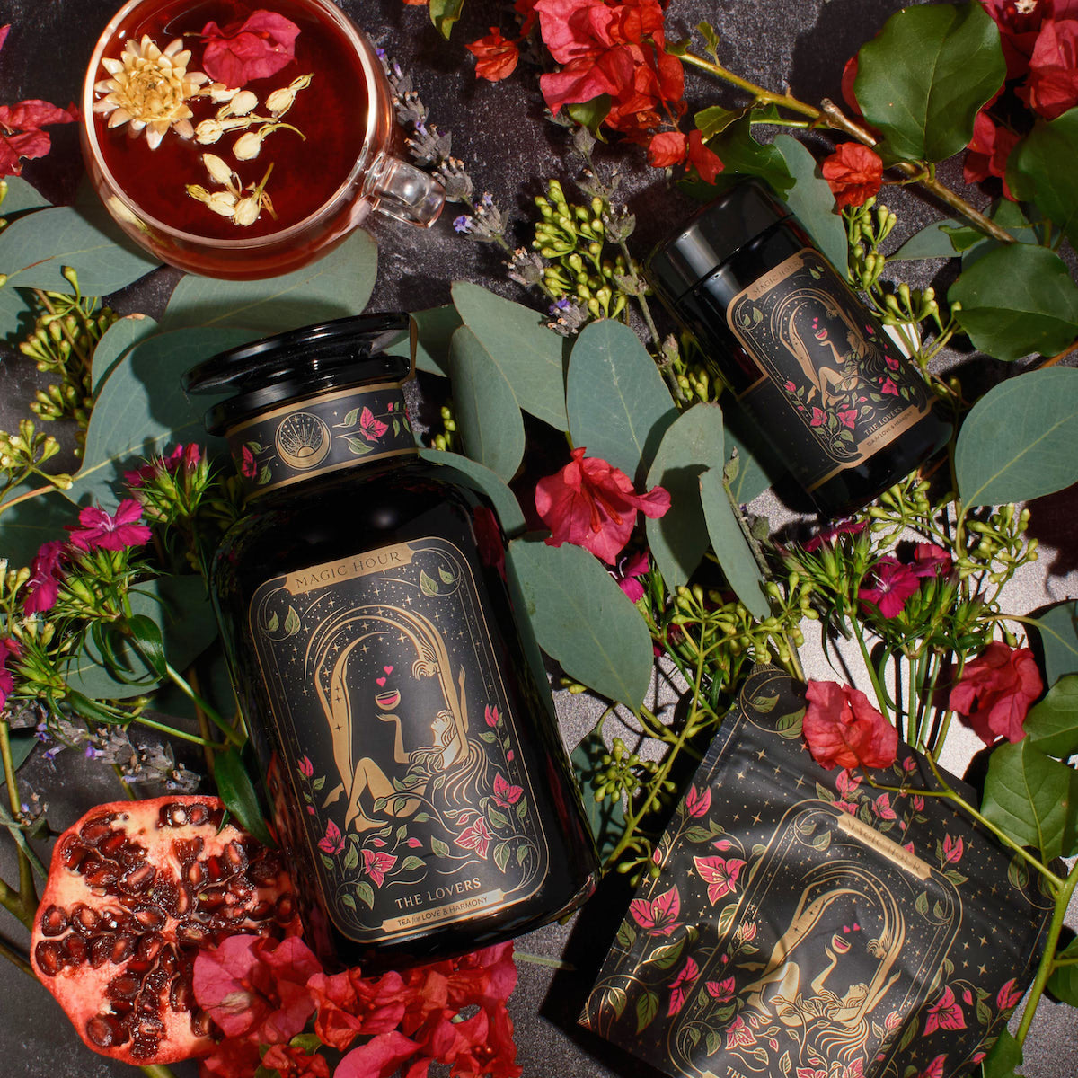 An ornate black jar labeled &quot;The Lovers&quot; from Magic Hour, surrounded by pink and red flowers, eucalyptus leaves, a pomegranate half, a tea cup with floating flowers, a smaller jar, and a box. The setting is a dark surface with a romantic and botanical theme featuring Organic Hibiscus.
