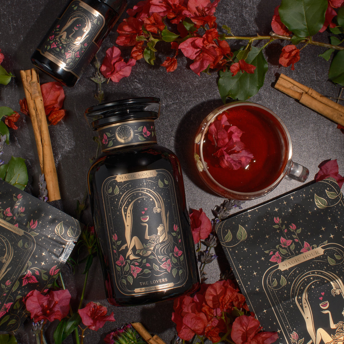 A stylized black bottle labeled "The Lovers" by Magic Hour is surrounded by red flowers, green leaves, cinnamon sticks, and a cup of organic hibiscus tea with flower petals. An accompanying box with mystical illustrations complements the bottle in a sophisticated, nature-themed flatlay.