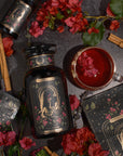 A stylized black bottle labeled "The Lovers" by Magic Hour is surrounded by red flowers, green leaves, cinnamon sticks, and a cup of organic hibiscus tea with flower petals. An accompanying box with mystical illustrations complements the bottle in a sophisticated, nature-themed flatlay.