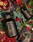 An ornate black jar labeled "The Lovers" from Magic Hour, surrounded by pink and red flowers, eucalyptus leaves, a pomegranate half, a tea cup with floating flowers, a smaller jar, and a box. The setting is a dark surface with a romantic and botanical theme featuring Organic Hibiscus.