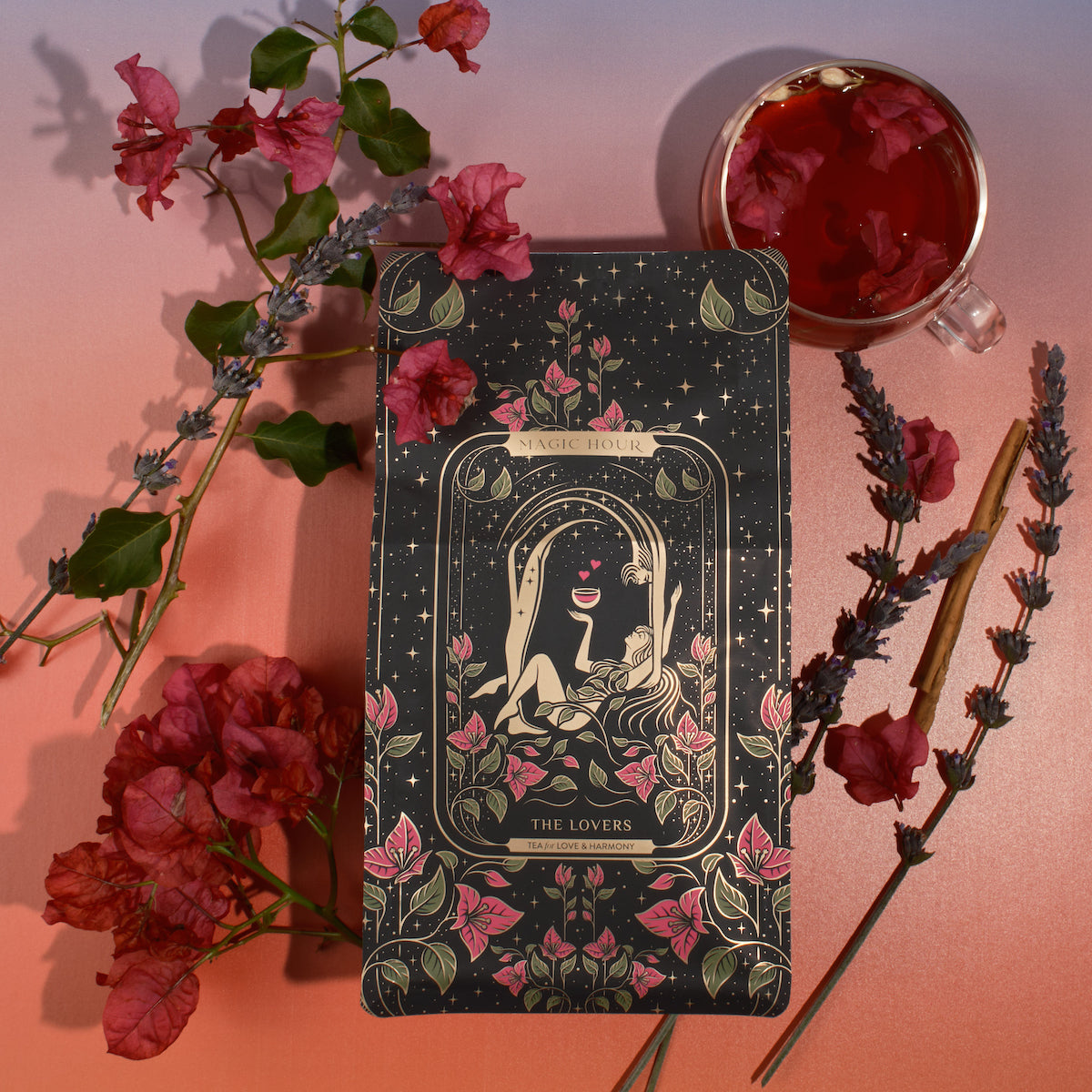 A tarot card titled &quot;The Lovers&quot; is surrounded by decorative flowers, lavender sprigs, and cinnamon sticks on a gradient background. The card features an illustration of two figures intertwined under a crescent moon and stars, with a chalice above them. A cup of Magic Hour&#39;s The Lovers tea infused with Organic Hibiscus sits nearby.