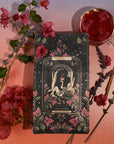 A tarot card titled "The Lovers" is surrounded by decorative flowers, lavender sprigs, and cinnamon sticks on a gradient background. The card features an illustration of two figures intertwined under a crescent moon and stars, with a chalice above them. A cup of Magic Hour's The Lovers tea infused with Organic Hibiscus sits nearby.