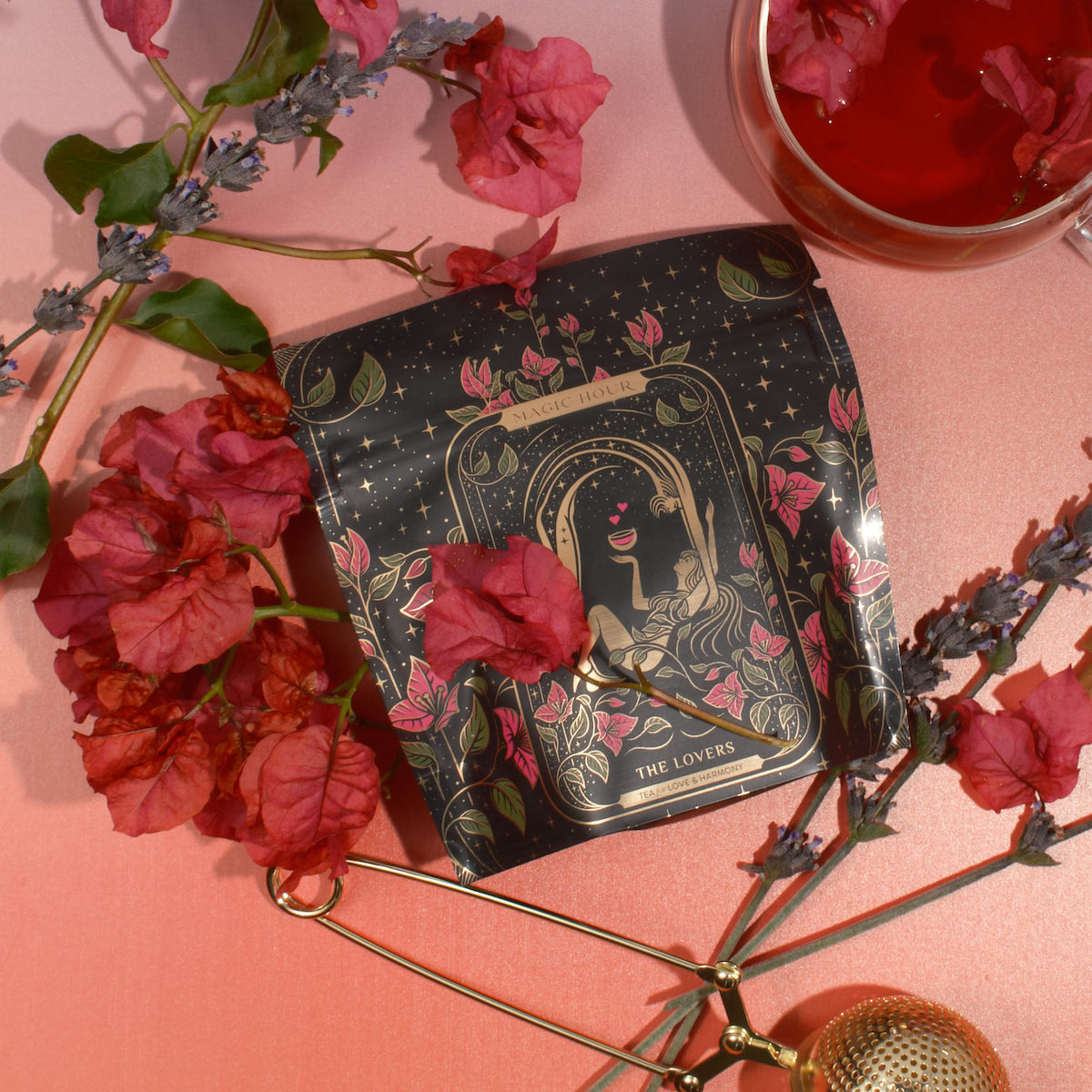 A packet of tea labeled &quot;The Lovers&quot; from Magic Hour is surrounded by red flowers and lavender sprigs on a gradient pink background. A golden tea infuser and a clear cup of red tea with Organic Hibiscus are also positioned nearby. The packet has an intricate, artistic design.
