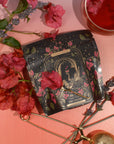 A beautifully designed tea package from the "12 Tarot Tea Sampler Bundle" rests on a pink surface, surrounded by red and lavender flowers. The tarot-inspired tea showcases an illustration of a hand holding a wine glass labeled "The Lovers," creating a Tarot Tasting Experience, with a nearby infuser.