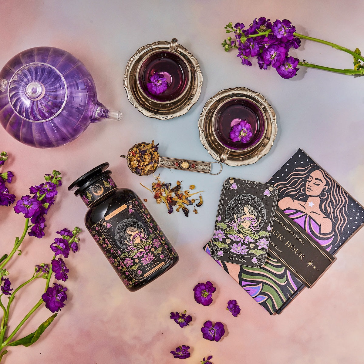 A vibrant flat lay featuring The Moon Box: a purple teapot and two teacups filled with herbal tea, a jar of Moon Blend loose tea leaves, a tea scoop, a deck of tarot cards, and an array of scattered flowers. This scene is rich with botanical and mystical elements, perfectly capturing the essence of the Magic Hour.