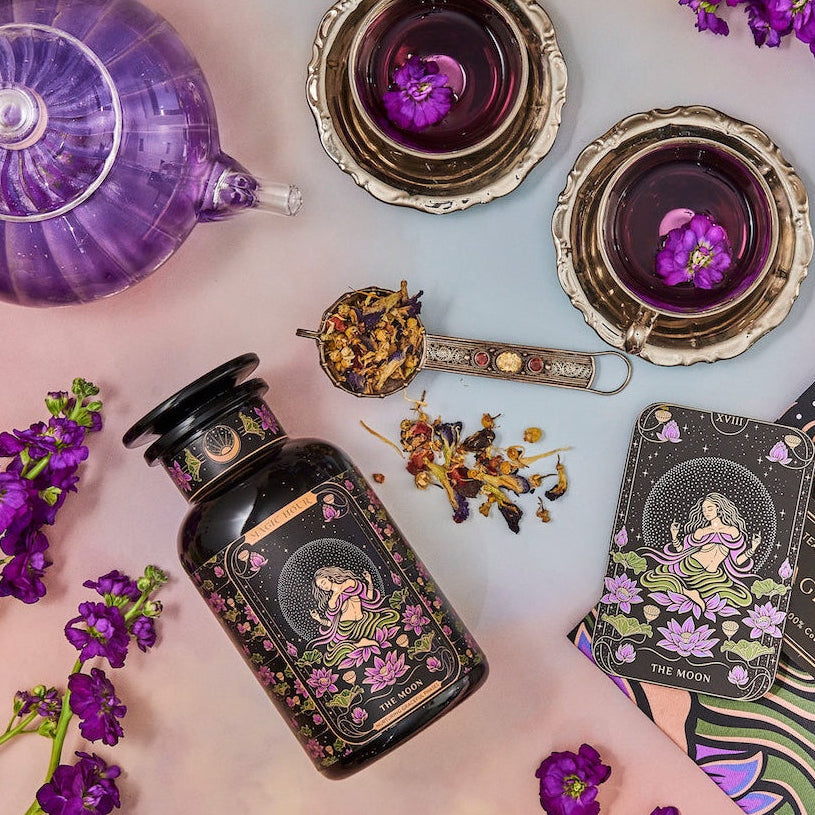 A colorful flat lay features a purple teapot and two teacups filled with herbal tea, surrounded by flowering purple plants. A jar of organic "The Moon - Tea for Nurturing Graceful Phases Case Packs" from Magic Hour, a tarot card labeled "The Moon," and a book titled "Magic Hour" complete the whimsical scene.
