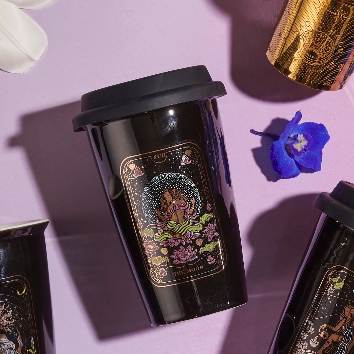 A black reusable cup with a mystical illustration featuring a meditative figure and lotus flowers, labeled &quot;The Queen of Cups - Tarot To Go!,&quot; is placed on a light pink surface. The lid is black, and nearby are another cup of Magic Hour Tea, a golden cylindrical object, a white flower petal, and a blue flower.