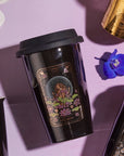 A black reusable cup with a mystical illustration featuring a meditative figure and lotus flowers, labeled "The Queen of Cups - Tarot To Go!," is placed on a light pink surface. The lid is black, and nearby are another cup of Magic Hour Tea, a golden cylindrical object, a white flower petal, and a blue flower.