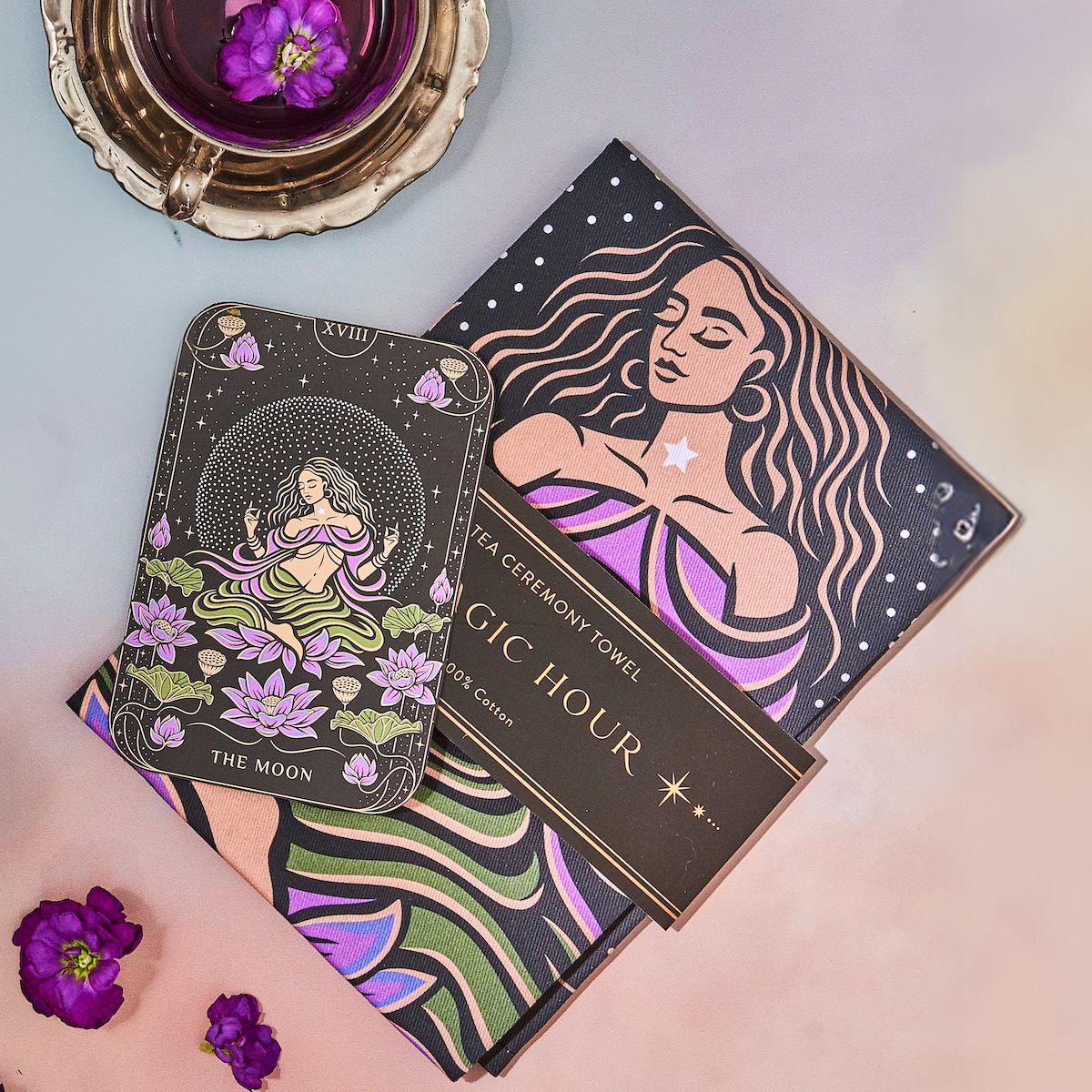 A tarot card depicting "The Moon" placed on top of a book titled "Magic Hour," featuring vibrant illustrations of a woman and celestial motifs. Nearby, purple flowers and a cup of The Moon - Tea for Nurturing Graceful Phases Case Packs from Magic Hour are set on a metallic tray, creating a mystical ambiance.