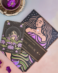 A tarot card depicting "The Moon" placed on top of a book titled "Magic Hour," featuring vibrant illustrations of a woman and celestial motifs. Nearby, purple flowers and a cup of The Moon - Tea for Nurturing Graceful Phases Case Packs from Magic Hour are set on a metallic tray, creating a mystical ambiance.