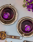 Two ornate silver cups brimming with a vibrant purple liquid, each adorned with a purple flower, rest on the table. Nearby, a metal tea infuser filled with dried herbs and Butterfly Pea Flower blooms, along with a small bunch of fresh purple flowers from Magic Hour's "The Moon - Tea for Nurturing Graceful Phases Case Packs," adds to the elegant scene.