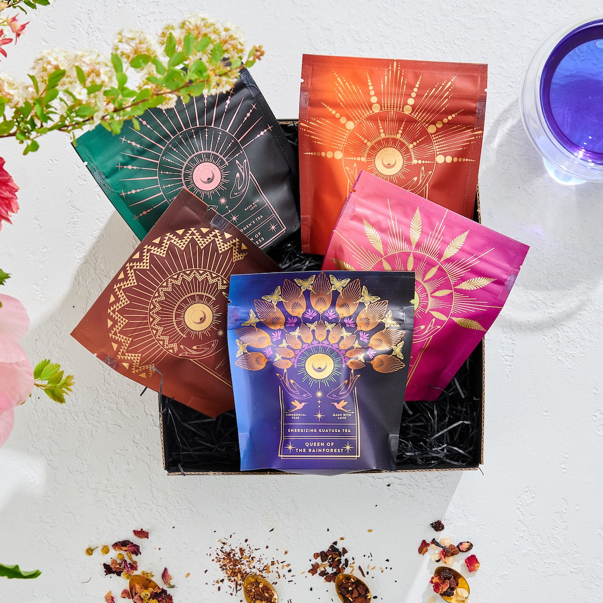 A box showcasing five vibrant packages from The Queen's Sampler Set features intricate designs. Flower petals and leaves surround the box, with a purple wellness tea glass on the side, set against a textured white backdrop.