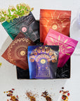 A box showcasing five vibrant packages from The Queen's Sampler Set features intricate designs. Flower petals and leaves surround the box, with a purple wellness tea glass on the side, set against a textured white backdrop.