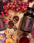 An artfully styled flat lay features a bottle labeled "Magic Hour," encircled by grapes, figs, raspberries, plums, and wellness tea. There's an array of tarot cards titled "The Queen of Cups" and assorted decorative items resting on the gold-patterned surface, crafting an enchanting scene perfect for the Monthly Magic First Sips Tea Subscription Box.