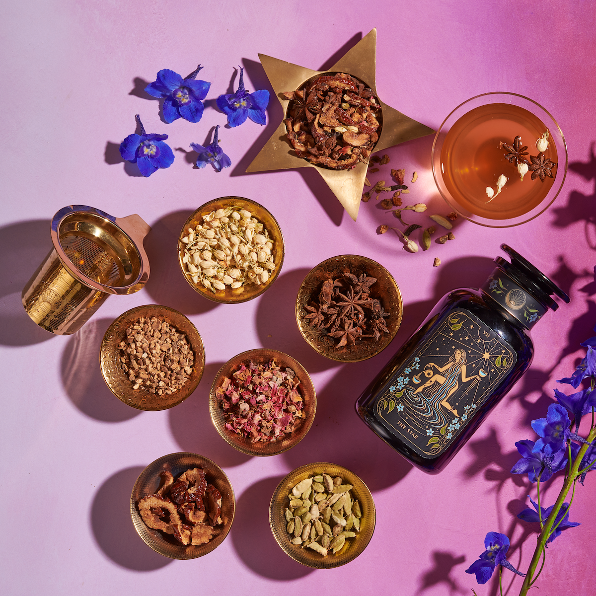 A flat lay featuring a variety of dried herbs and spices in small bowls, a glass teacup with star anise, a gold strainer, a large dark bottle with floral design displaying "LE 5PM," and blue flowers on a pink and purple gradient background. Perfect for enjoying the Monthly Magic 3-Month Tea Subscription Box from Magic Hour.