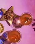 Two glasses of orange-colored "The Star: Vanilla-Ginger Beauty Potion with Jasmine & Shatavari" from Magic Hour, garnished with star anise flowers and other dried botanicals, placed on a pink background. A gold star-shaped container filled with mixed dried botanicals and a small bowl of herbs are nearby. Blue and purple flowers are scattered around.