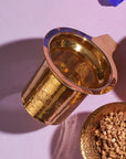 A gold tea infuser with fine mesh sits on a light pink surface, casting a shadow. Nearby, a small bowl filled with ginger pieces is partially visible on the right, hinting at the preparation of an exquisite Beauty Potion from The Star Box. The overall setting suggests a detailed tea preparation scene.