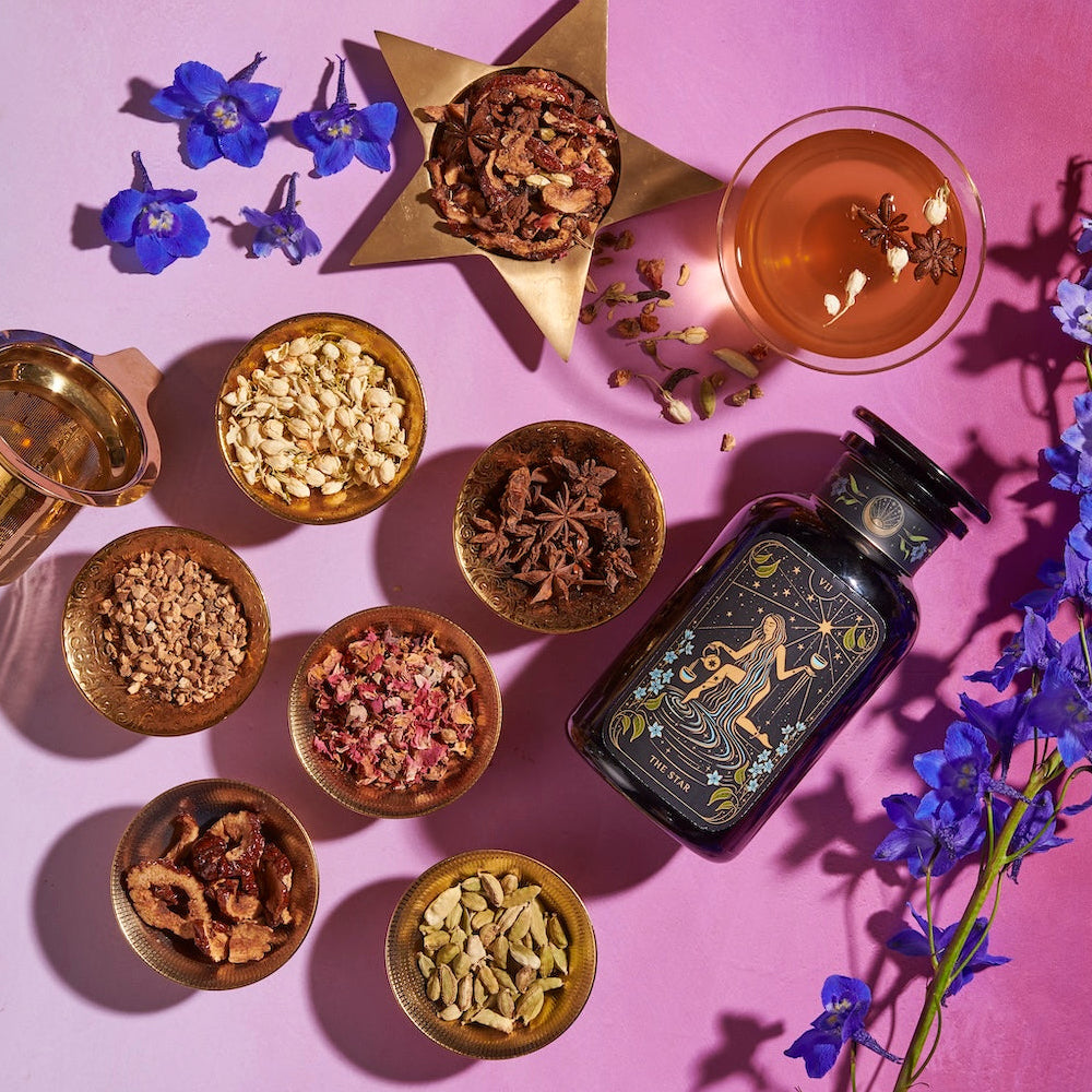 A flat lay of The Star Box items, including assorted spices and ingredients arranged in small bowls on a pink surface. It features an artisan tea strainer, an artistically designed bottle, a glass of the Star Vanilla Ginger Beauty Potion, a star-shaped bowl of dried herbs, and scattered blue flowers.