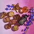A flat lay of The Star Box items, including assorted spices and ingredients arranged in small bowls on a pink surface. It features an artisan tea strainer, an artistically designed bottle, a glass of the Star Vanilla Ginger Beauty Potion, a star-shaped bowl of dried herbs, and scattered blue flowers.